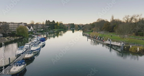 Obraz London, Teddington: Calm waters of River Thames reflecting sky and trees, with boats moored on left bank and a pier on the right bank. Aerial drone flight over residential calm area in capital of UK