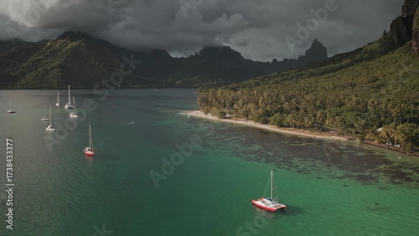 Obraz Stunning aerial view of yachts in turquoise lagoon bay, tropical island beach in French Polynesia, green mountains and dramatic cloudy sky. Remote wild nature paradise, exotic summer travel background