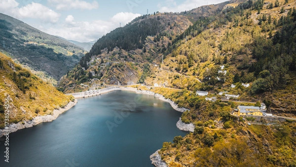 Fototapeta A serene mountain reservoir between steep forested slopes, concrete dam, and hillside houses with winding road under soft clouds, clear teal water, and rugged rocky shoreline