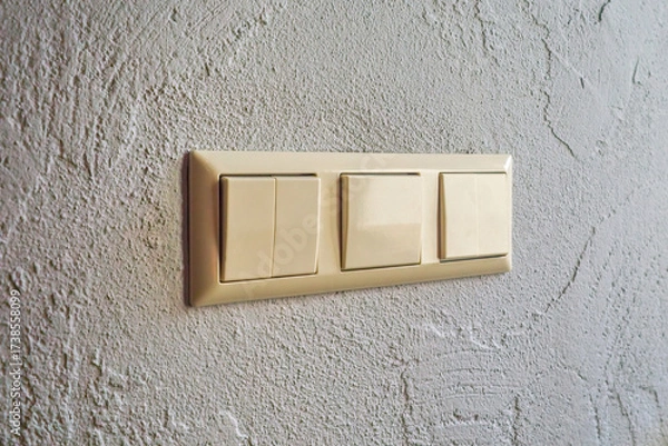 Fototapeta Triple beige light switch mounted on a textured wall indoors. Electrical switch panel for controlling lighting.