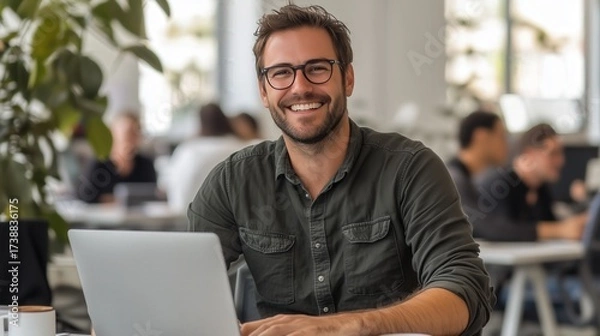 Fototapeta A casual young man smiles while working on his laptop at a table, enjoying coffee and productivity.