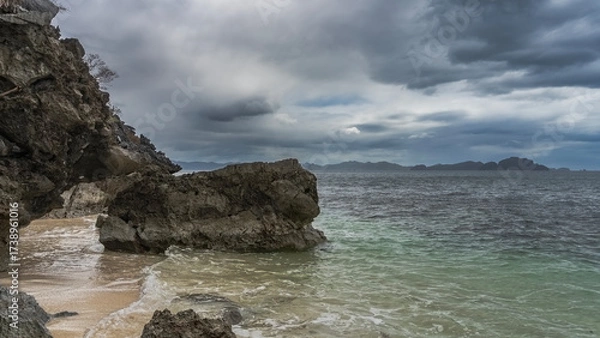 Fototapeta Picturesque boulders in the ocean. Coastal cliffs overhang the water. Sea waves are foaming on a sandy beach. There are no people. Mountains on the horizon. Clouds in the sky. Philippines. Palawan