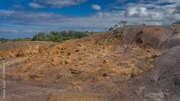 Fototapeta A unique geological landmark. 23 coloured earth of Mauritius. Multi-colored sand hill against a background of blue sky and clouds. Green vegetation in the distance. La Vallée Des Couleurs Nature Park