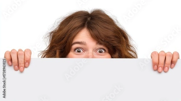 Fototapeta A surprised or curious young woman with wide eyes and curly brown hair is peeking over a blank white sign or billboard, holding its edges with her hands.