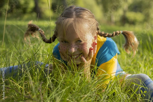 Obraz smiling blond girl with funny pigtail sits in grass. She is wearing a yellow T-shirt and a blue sarafan, and she has freckles on her nose.