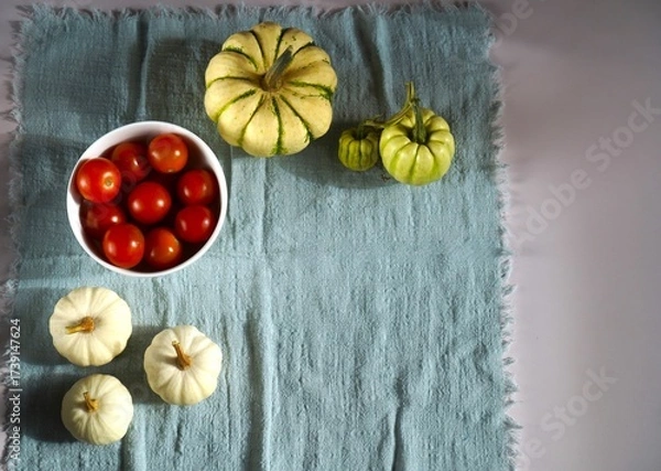 Obraz op view of ripe red cherry tomatoes in a white bowl alongside assorted gourds placed artistically on a textured blue fabric. The image highlights autumn harvest and organic produce.