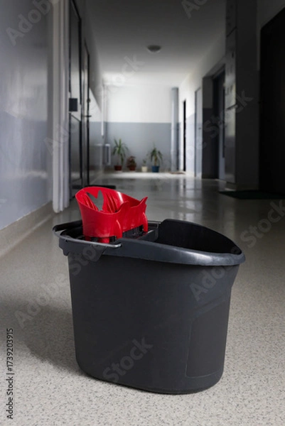 Fototapeta Black cleaning bucket with a red mop resting inside, placed in a hallway during daytime