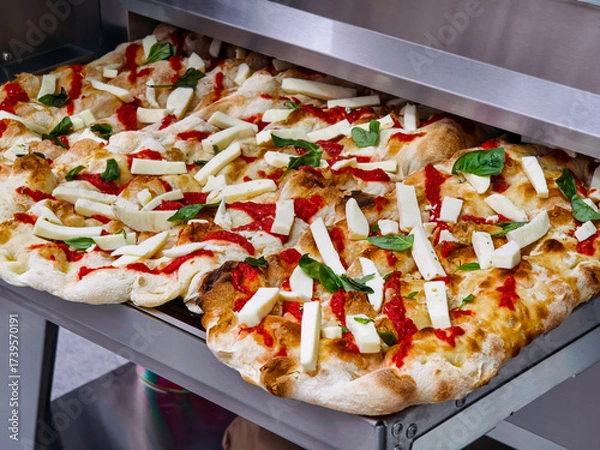 Fototapeta Freshly baked pizza with tomato sauce and mozzarella cheese, topped with basil, ready to serve in a busy kitchen