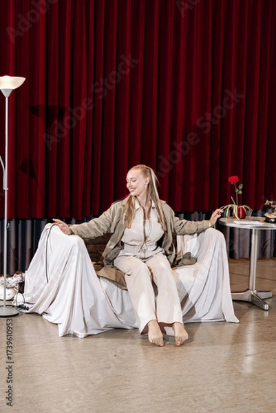 Fototapeta A beautiful blonde sits on a chair in a room with red curtains. Theater stage.