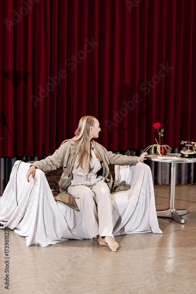 Fototapeta A beautiful blonde sits on a chair in a room with red curtains. Theater stage.