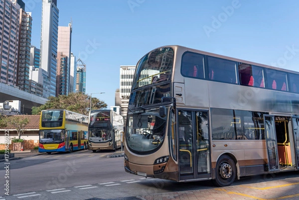 Fototapeta Double-decker buses are positioned at a busy city intersection, with one bus open for passengers while others wait. The skyline features modern buildings under clear skies