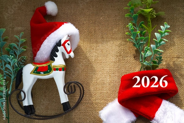 Obraz  Festive image of a Santa hat placed on a wooden rocking horse, accompanied by vibrant greenery and the year 2026. Perfect for holiday and New Year-themed designs and decorations
