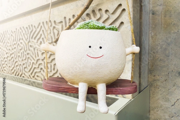 Fototapeta Charming planter with smiling face is hanging on swing, holding vibrant green plants. Playful decor, modern urban environment. Sunlight warm glow, delightful scene of indoor gardening.