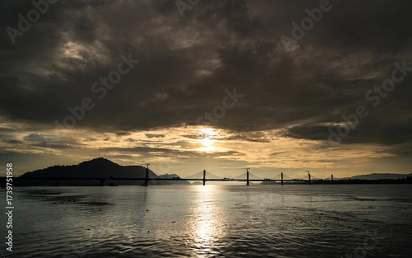 Fototapeta A scenic view of a cable-stayed bridge over the Brahmaputra River during sunset, with reflections shimmering on the water and dramatic clouds in the sky