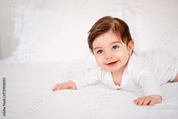 Obraz A baby under a blanket on a white bed at home, a place for text, a cute smiling little child getting ready for bed