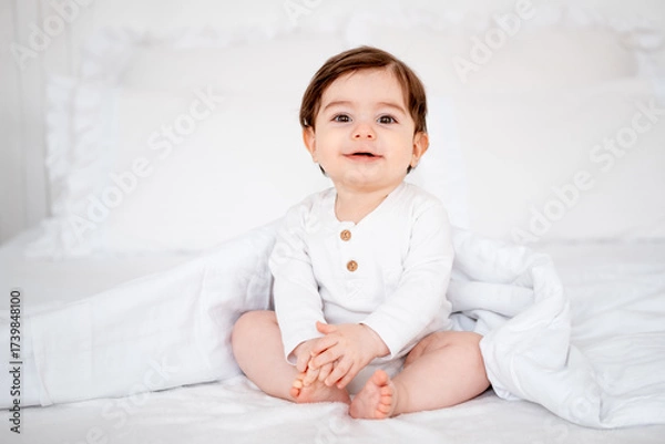 Obraz A baby under a blanket on a white bed at home, a place for text, a cute smiling little child getting ready for bed