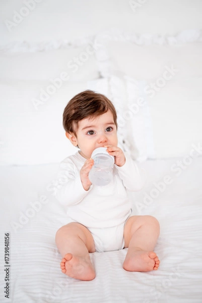 Obraz baby with a bottle of milk or water on a white bed at home, baby food feeding concept, place for text, cute smiling little baby