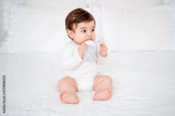 Obraz baby with a bottle of milk or water on a white bed at home, baby food feeding concept, place for text, cute smiling little baby