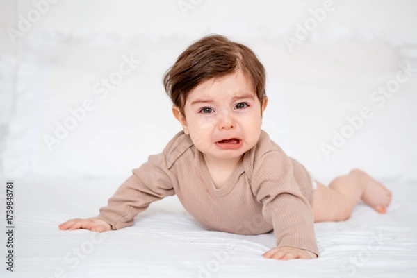 Obraz A crying baby on a white bed at home, a place for text