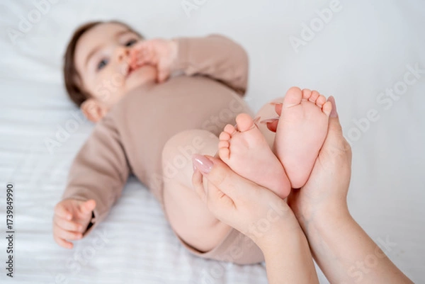Obraz baby's legs on a white bed, a place for text, mom's hands holding the baby's legs