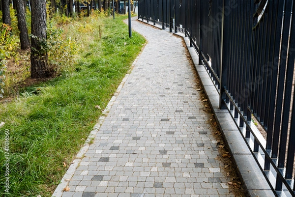 Fototapeta A beautifully curving paved pathway runs through a vibrant landscape, surrounded by lush greenery and a charming fence that enhances the picturesque scenery of this tranquil outdoor space