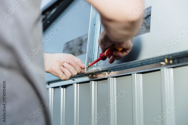 Fototapeta A man uses a screwdriver to attach a roof part while installing a metal shed.