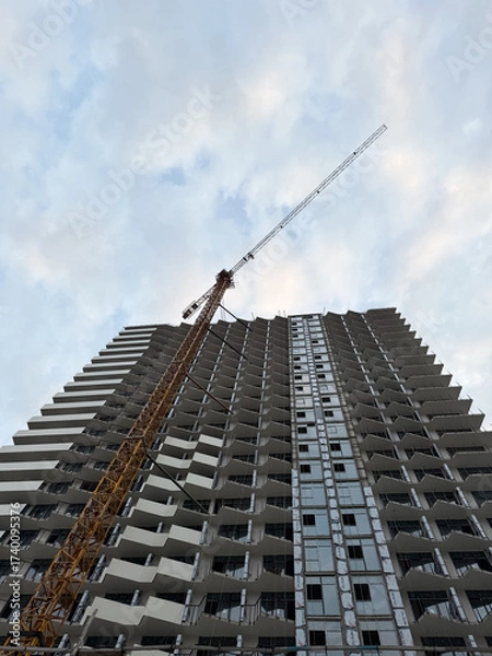 Fototapeta High-rise building under construction with a crane on the side, showing incomplete exterior and open windows, photographed from below