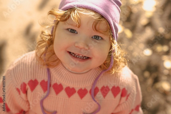 Fototapeta A cheerful toddler with curly hair and a purple hat smiles brightly outside, surrounded by a warm, sunny atmosphere and golden leaves in the background.