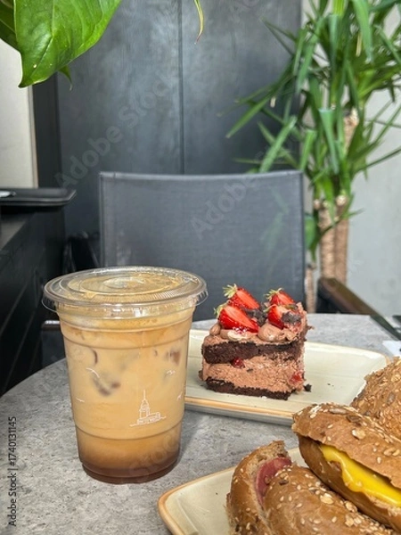 Fototapeta An iced coffee in a plastic cup, a chocolate strawberry cake slice, and two whole grain sandwiches with cheese on a round gray table, indoors.