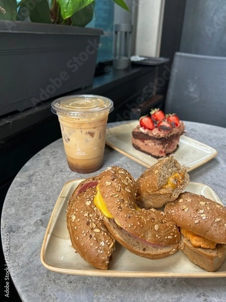 Fototapeta A table with iced coffee, chocolate cake with strawberries, and sesame bagels filled with cheese and meat. Flowerpot and chair are partially visible.