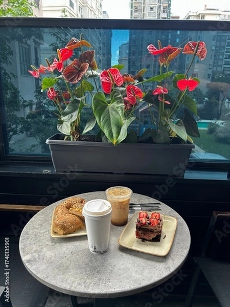 Fototapeta A small round table on a balcony holds two drinks, two bagels, and a slice of chocolate cake with strawberries, with red flowers and buildings in the background.
