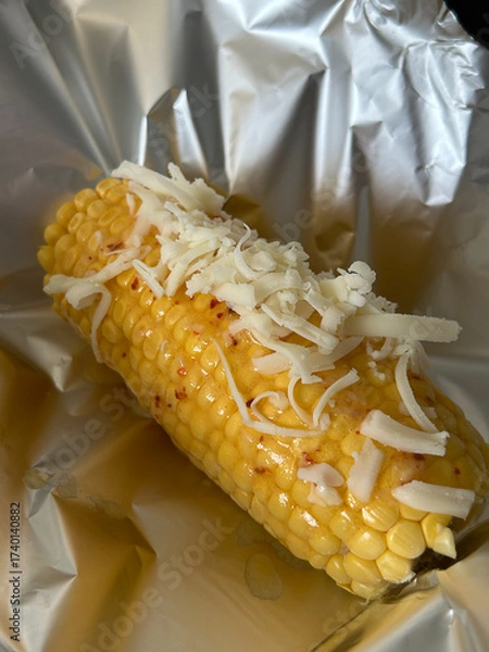 Fototapeta Close-up of a cooked corn cob topped with grated cheese, placed on aluminum foil. The corn appears to be buttered and mildly spiced
