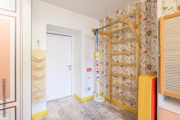 Fototapeta A child's playroom or hallway with a wooden climbing frame against bird-themed wallpaper, a rope ladder, and woven storage. A white door and wood-style flooring complete the bright, playful space