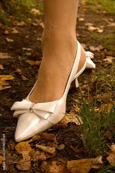 Obraz A woman’s foot in a white shoe with a bow and low heel stands on the ground covered with dry autumn leaves and grass in a park.