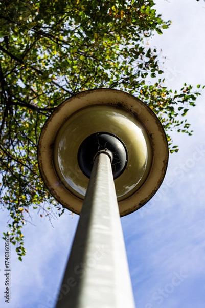 Fototapeta Extreme low-angle view of a vintage yellow street lamp and pole stretching into a bright blue sky, with green tree leaves framing the top, showcasing nature and urban contrast.

