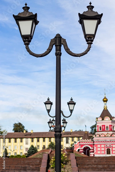 Fototapeta A classic twin lamppost stands tall in the foreground against a historical cityscape with an ornate church and buildings under a bright blue, cloudy sky, suggesting a public square.

