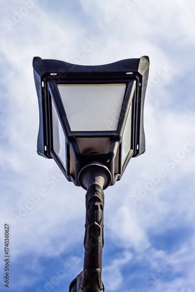Fototapeta A low-angle shot of a classic black street lamp (lantern) against a bright blue sky with wispy clouds. The design suggests vintage or historical urban architecture.

