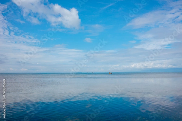 Fototapeta A wide view of the calm ocean stretches under a bright blue sky with scattered clouds on the horizon. The scene captures tranquility, open space, and peaceful seascape beauty.