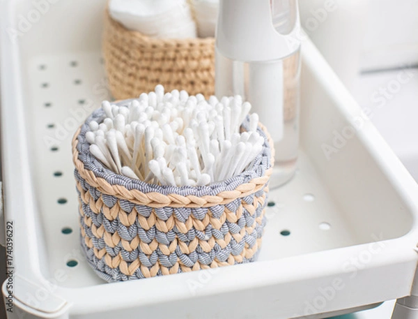 Fototapeta Woven basket filled with cotton swabs sits on a bathroom tray alongside a spray bottle and additional storage, showcasing organized personal care essentials