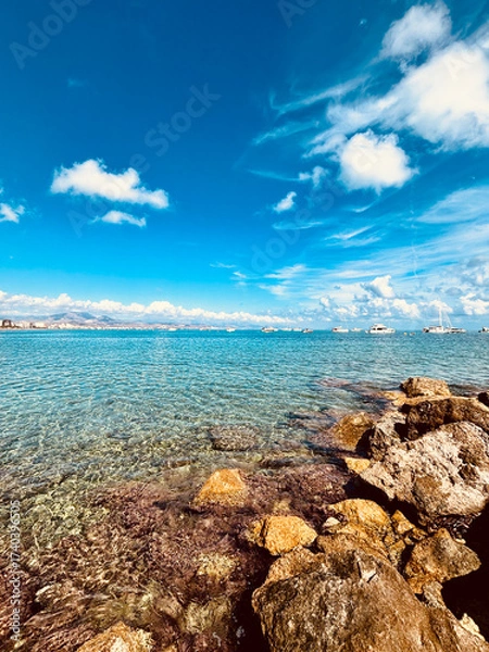 Fototapeta Clear turquoise water gently laps against rocky shoreline under a bright blue sky with fluffy clouds, creating a serene coastal landscape perfect for relaxation and escape