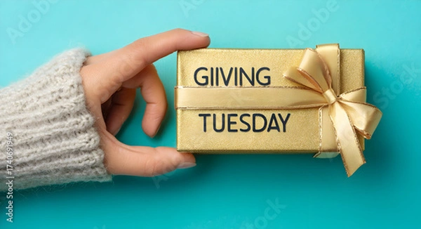 Fototapeta Hand holding a gold giving tuesday gift box with a gold ribbon on a blue background overhead shot