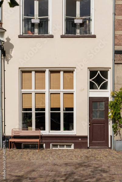 Fototapeta Charming building facade with a bench, textured windows, and brick pavement in soft sunlight.