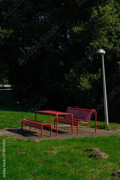Fototapeta Red metal picnic set on a concrete pad by green grass and a sleek silver lamp post.