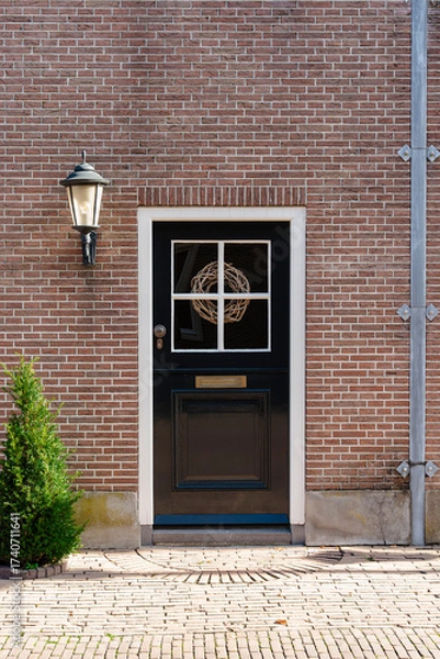 Fototapeta Elegant door with a wreath on a brick building, complemented by a lamp and greenery on a brick path.