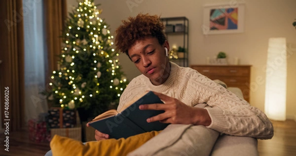 Fototapeta A boy is listening to music through wireless headphones while reading an interesting book, turning the pages and fully giving himself away to the story on a winter evening by the Christmas tree.