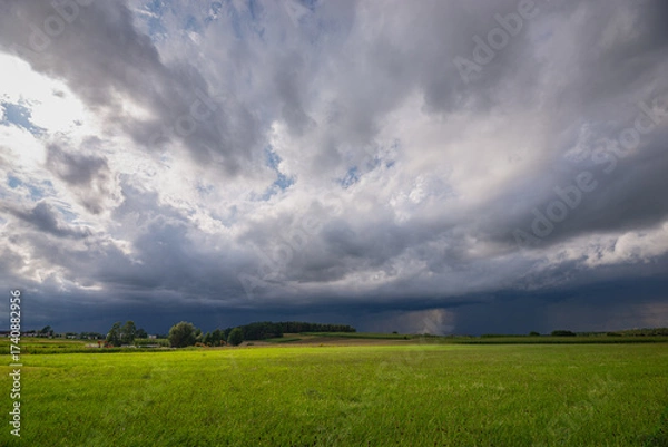 Fototapeta Stormy skies above an open green field, dramatic weather, clouds and expansive rural landscape.