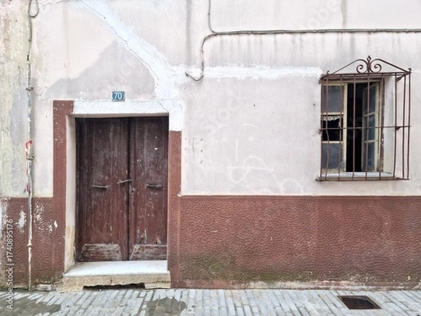 Fototapeta Old weathered wooden door and window with rusty bars on aged wall of abandoned house