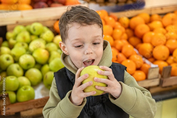 Fototapeta Close up happy little boy eating green apple at farmer's market with apple and tangerine counter in the background. Childhood and healthy nutrition concept. High quality photo