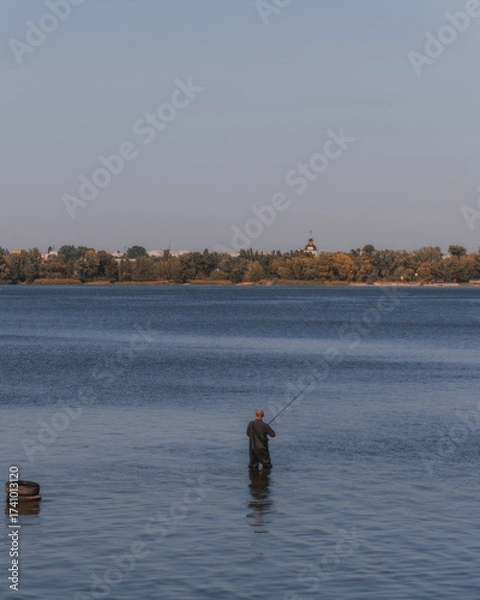 Fototapeta A lone fisherman stands knee-deep in the water of a large body of water, fishing in the morning. A figure in wading gear is positioned at the bottom of the frame, conveying a sense of calm.