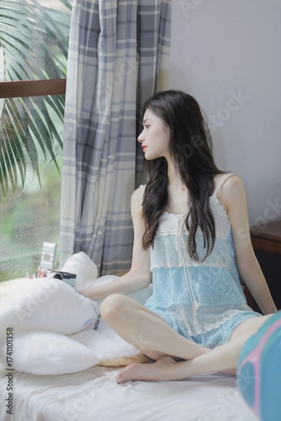 Fototapeta Woman in light blue lace dress sitting by the window with a soft toy, expressing a calm and thoughtful mood.